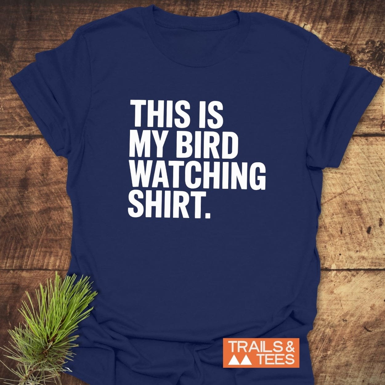 Navy-blue Bird Watching T-Shirt with bold white text, laid flat. Features durable print, taped shoulders, and a rustic pine sprig accent.