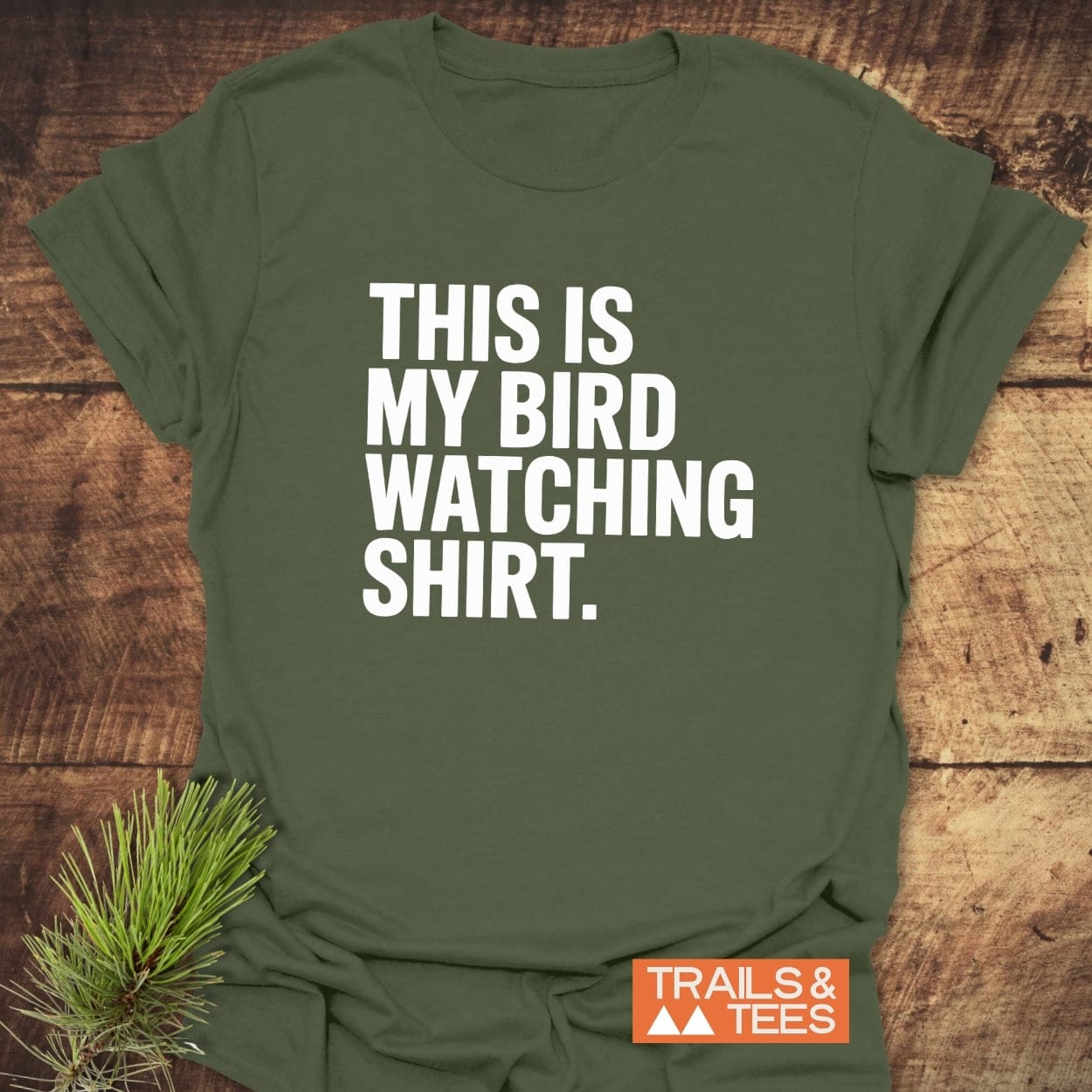 Bird Watching T-Shirt on wooden surface; features THIS IS MY BIRD WATCHING SHIRT text, orange “TRAILS & TEES” logo, and a pine sprig accent.