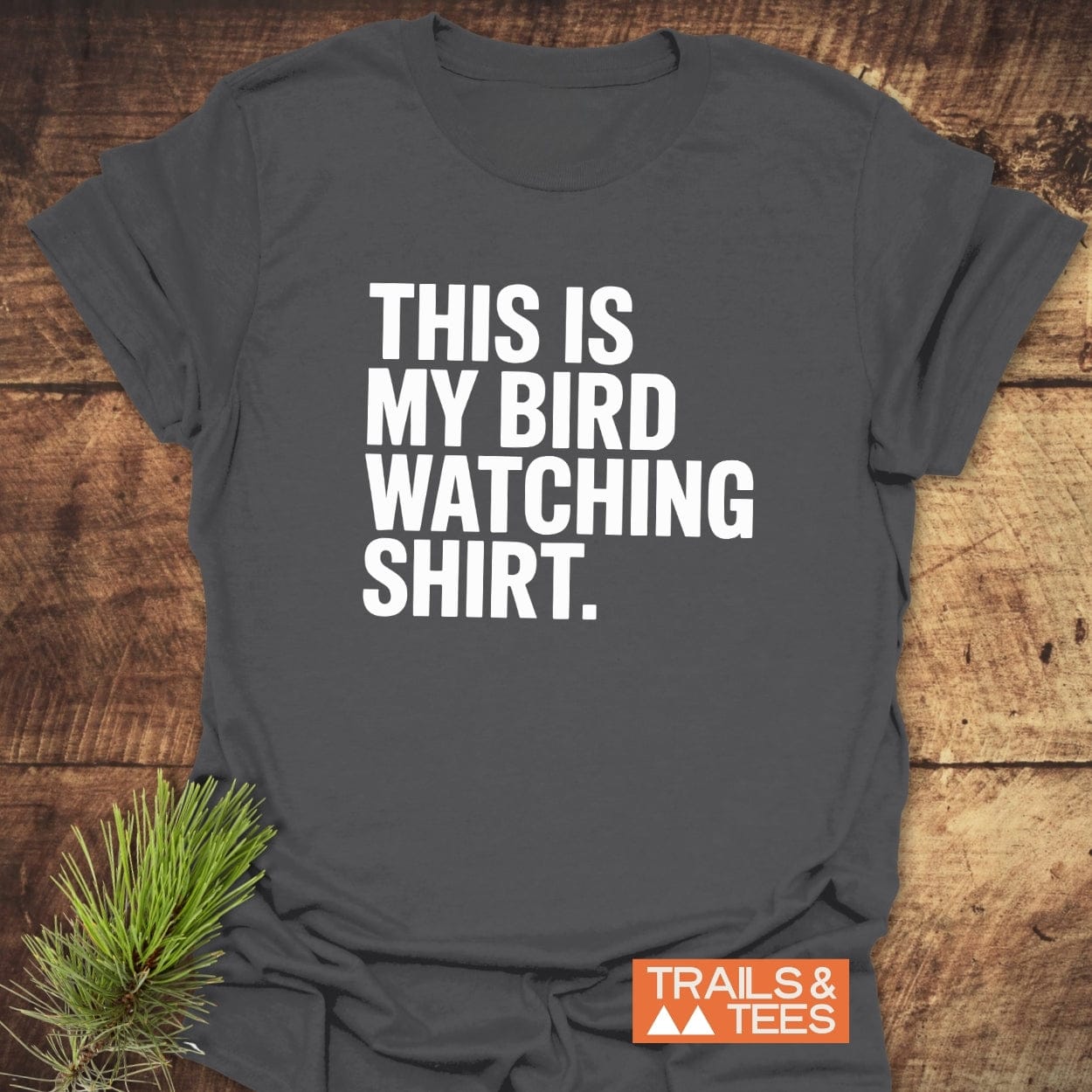Bird Watching T-Shirt on wood, featuring bold white slogan. Orange TRAILS & TEES logo visible. Pine sprig beside shirt.