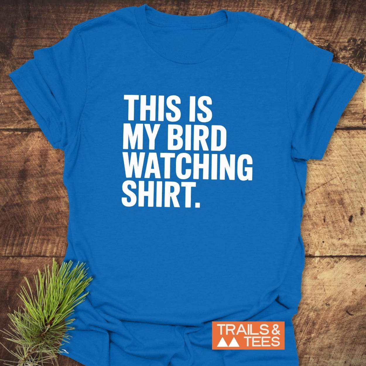Bird Watching T-Shirt on rustic wood, features THIS IS MY BIRD WATCHING SHIRT in white. Includes TRAILS & TEES logo with pine needles for outdoor appeal.