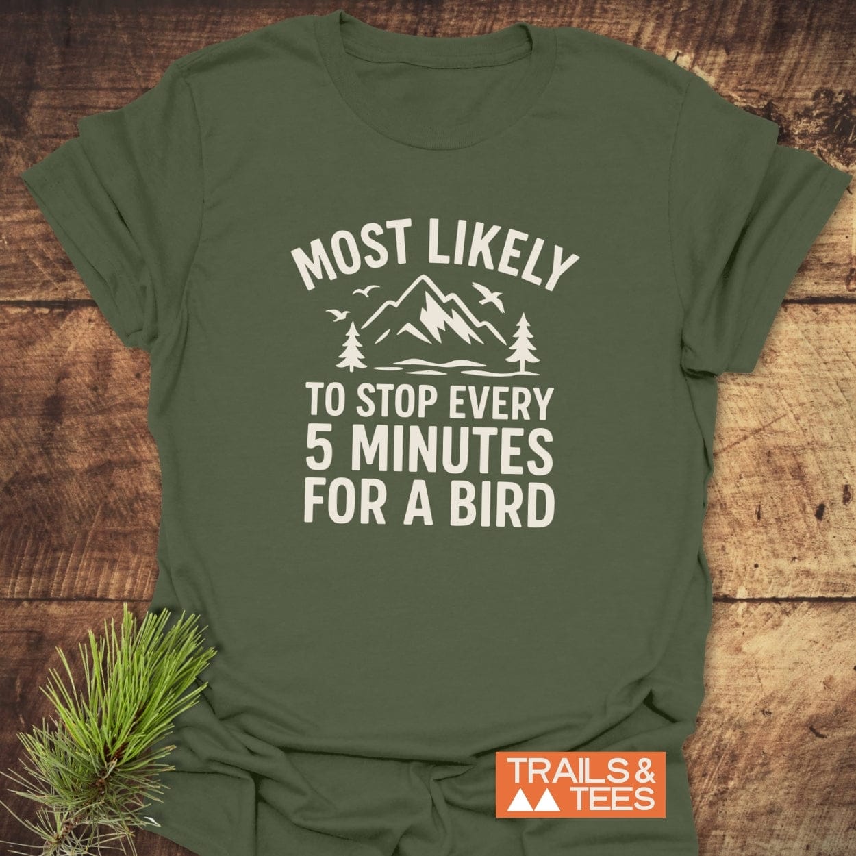 Most Likely Bird Watcher T-Shirt features a mountain scene print, slogan, and orange label, showcasing its outdoor-inspired design on a wooden surface.