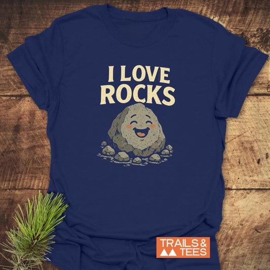 I Love Rocks T-Shirt on rustic wood, featuring a cartoon rock with pebbles. Includes Trails & Tees logo and pine branch for a nature-inspired touch.