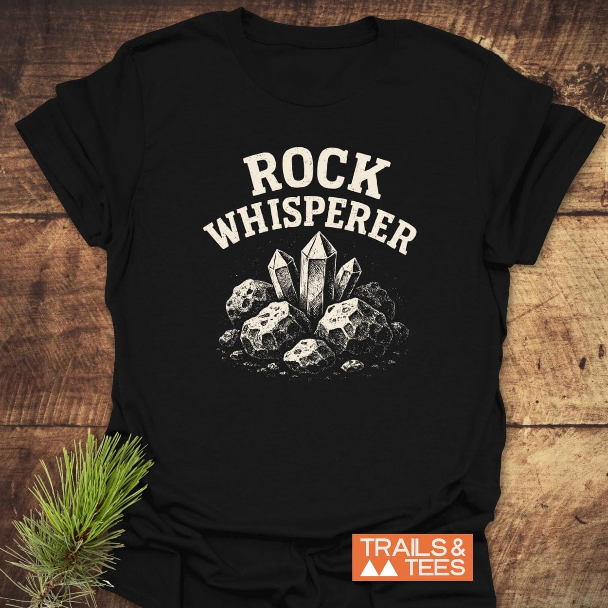 Rock Whisperer T-Shirt featuring white print of rocks and crystals, orange “TRAILS & TEES” label, styled with pine sprig on wooden surface.