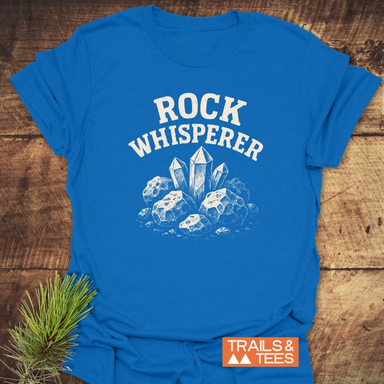Rock Whisperer T-Shirt featuring a white crystal graphic and ROCK WHISPERER text, laid on wood with the TRAILS & TEES logo and pine needles.