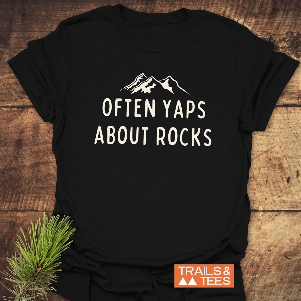 Often Yaps About Rocks T-Shirt featuring a mountain line-drawing and text, laid flat on wood. Includes Trails & Tees logo, showcasing its outdoor adventure theme.