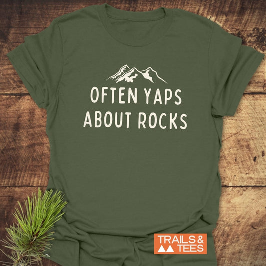 Often Yaps About Rocks T-Shirt on wooden surface, showcasing white mountain silhouette and text. Features durable print, soft ring-spun cotton, and eco-friendly production.