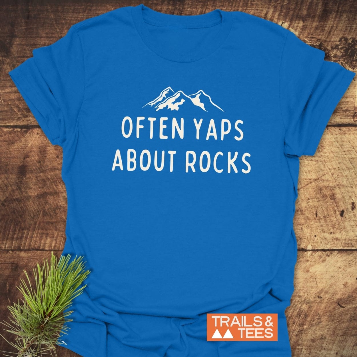 Often Yaps About Rocks T-Shirt with mountain silhouette, crafted from soft cotton for comfort. Features durable print, orange label, and taped shoulders for lasting shape.