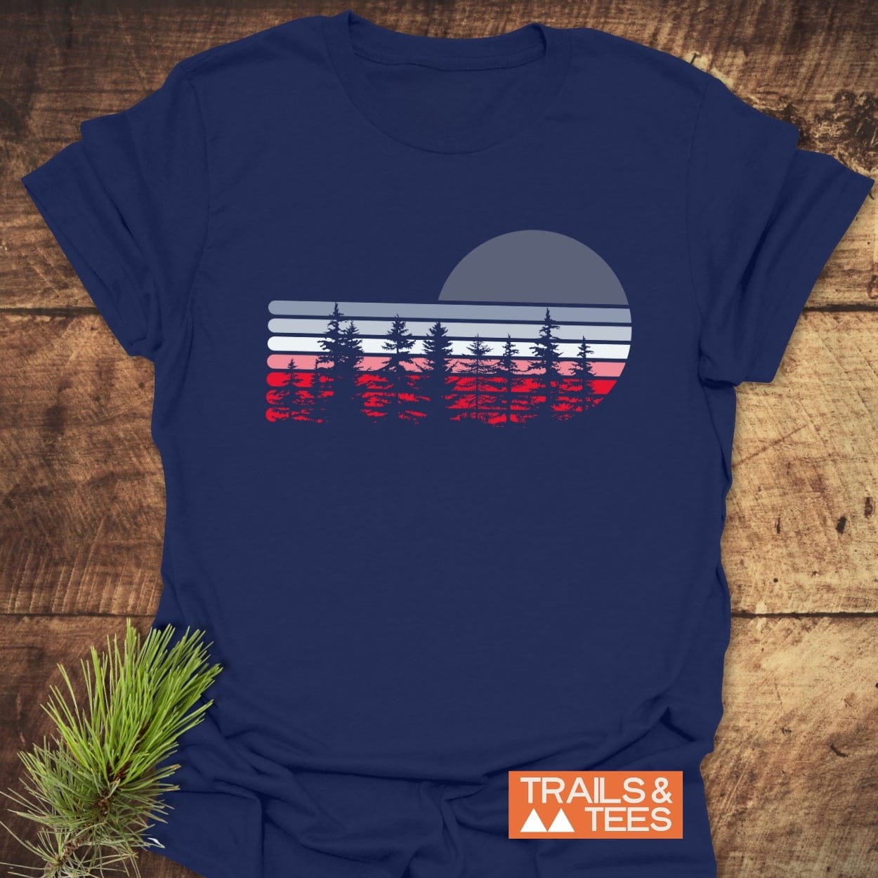 Retro Sunset Pines T-Shirt laid on wood, featuring a retro landscape graphic with pine silhouettes. Includes “TRAILS & TEES” logo and pine sprig for styling.
