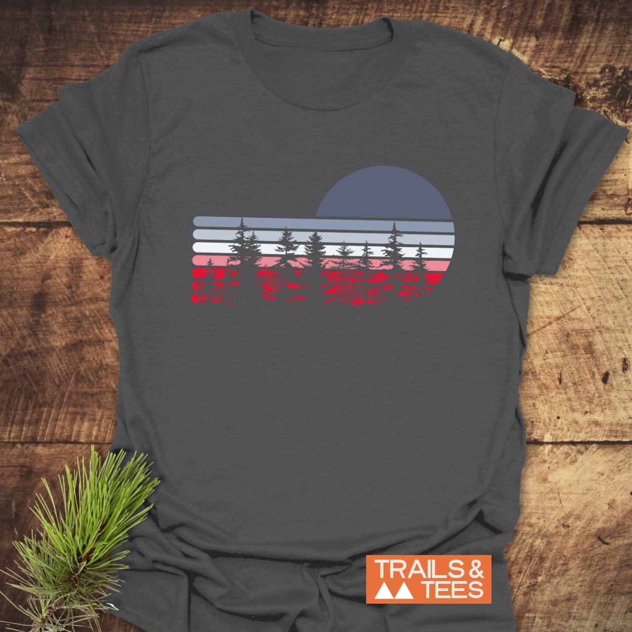 Retro Sunset Pines T-Shirt featuring a retro graphic with stripes, pine trees, and sun/moon, emphasizing outdoor adventure. Includes TRAILS & TEES logo and evergreen sprig.