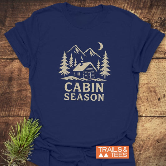 Cabin Season T-Shirt featuring a rustic cabin scene with pine trees and mountains, screen-printed on soft, lightweight cotton, styled with a pine sprig.