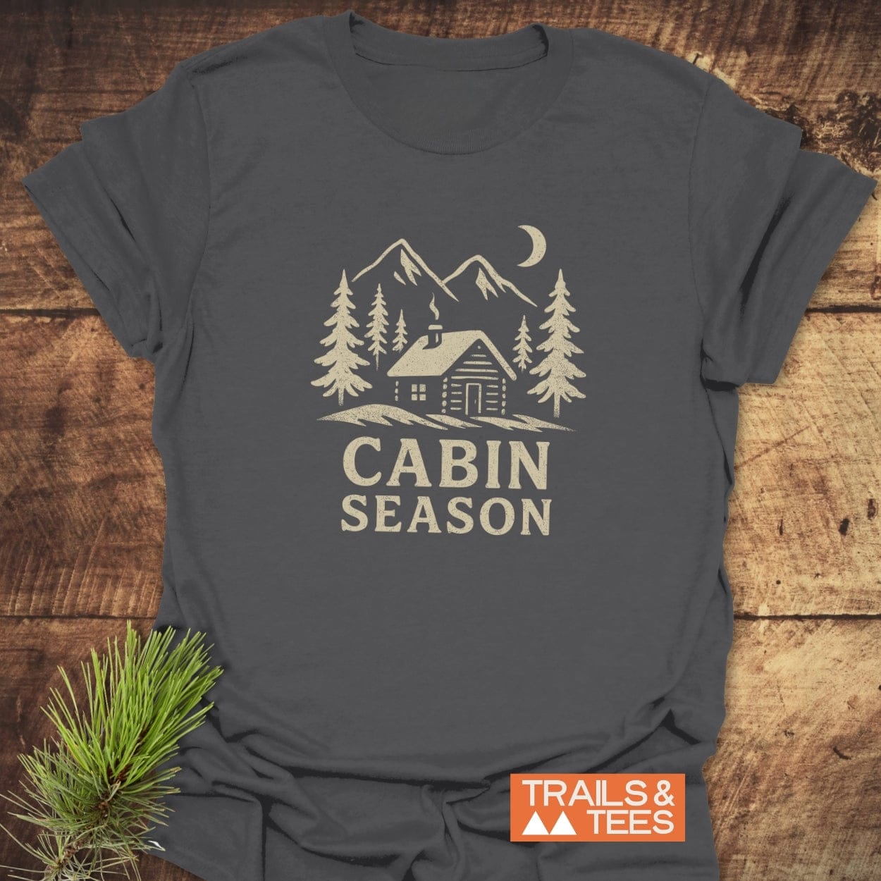 Cabin Season T-Shirt with mountain cabin graphic, pine trees, crescent moon, and TRAILS & TEES logo, showcasing adventure-ready, soft ring-spun cotton.