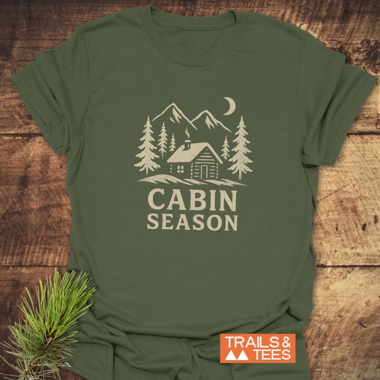 Cabin Season T-Shirt featuring a rustic log cabin graphic, pine trees, mountains, and moon, representing adventure and outdoor spirit. Includes Trails & Tees logo.