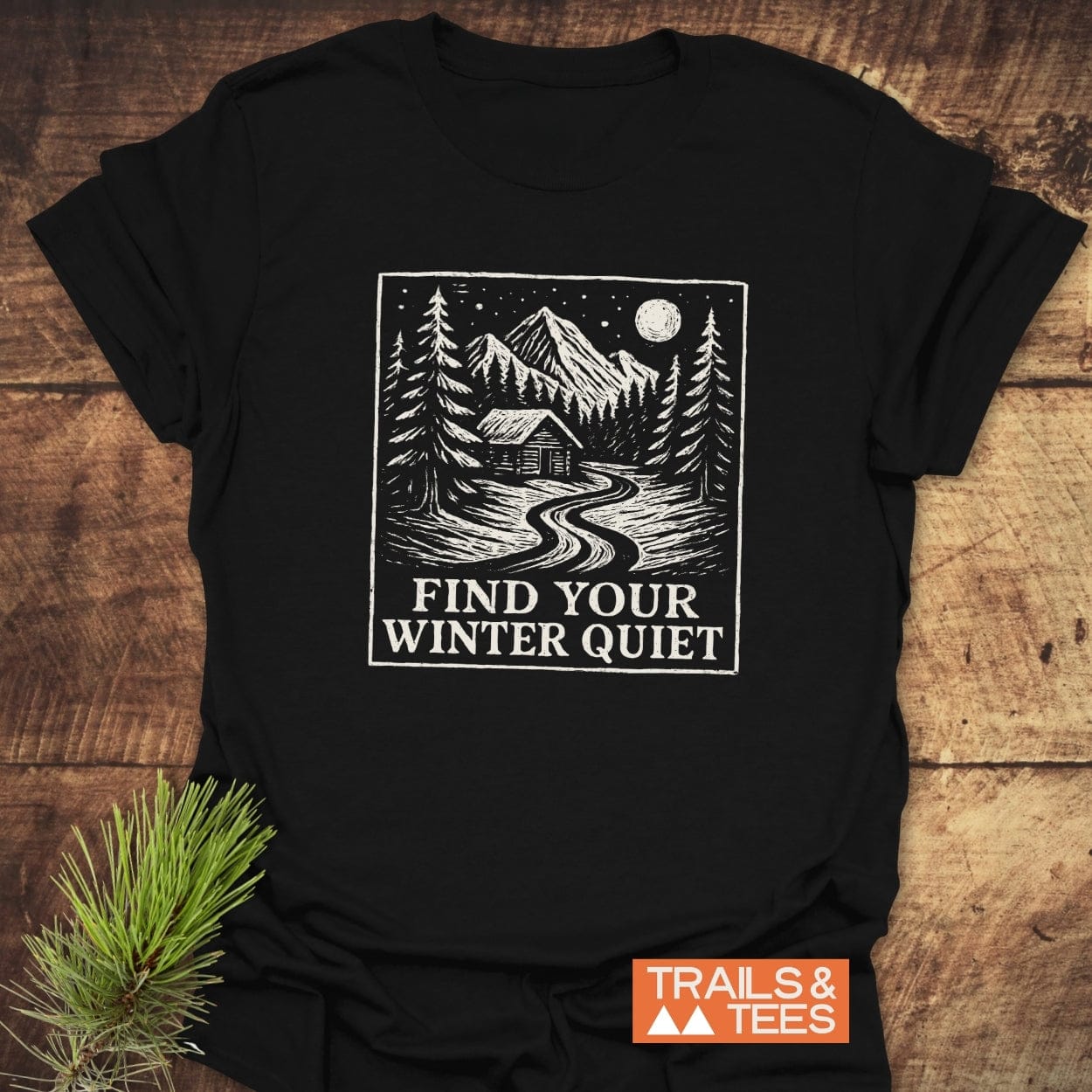 Find Your Winter Quiet Cabin T-Shirt: black tee featuring a snowy mountain scene graphic and slogan, with orange TRAILS & TEES logo, beside a pine sprig.