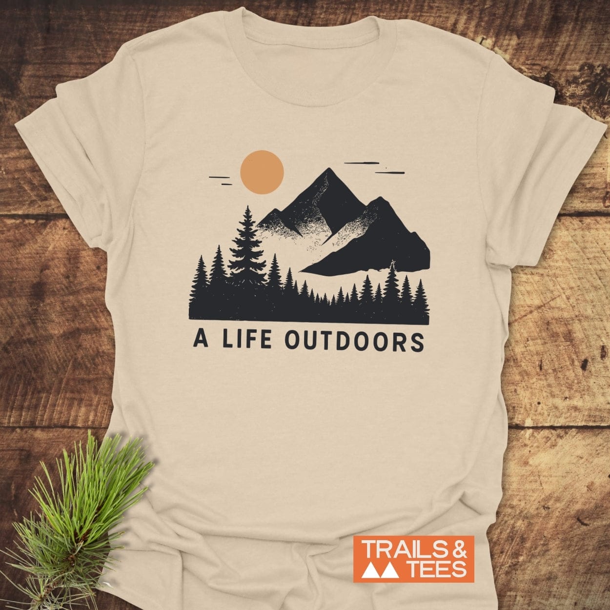 A Life Outdoors T-Shirt with a mountain and pine graphic, slogan, and Trails & Tees badge, laid on wood with pine sprigs.