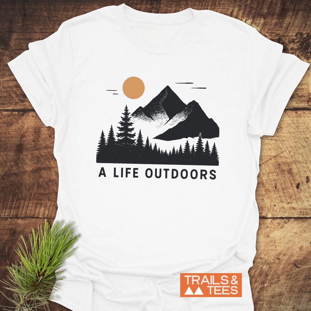 A Life Outdoors T-Shirt with mountain and pine tree graphic, orange sun, and slogan. Made of soft, lightweight cotton, featuring durable prints.