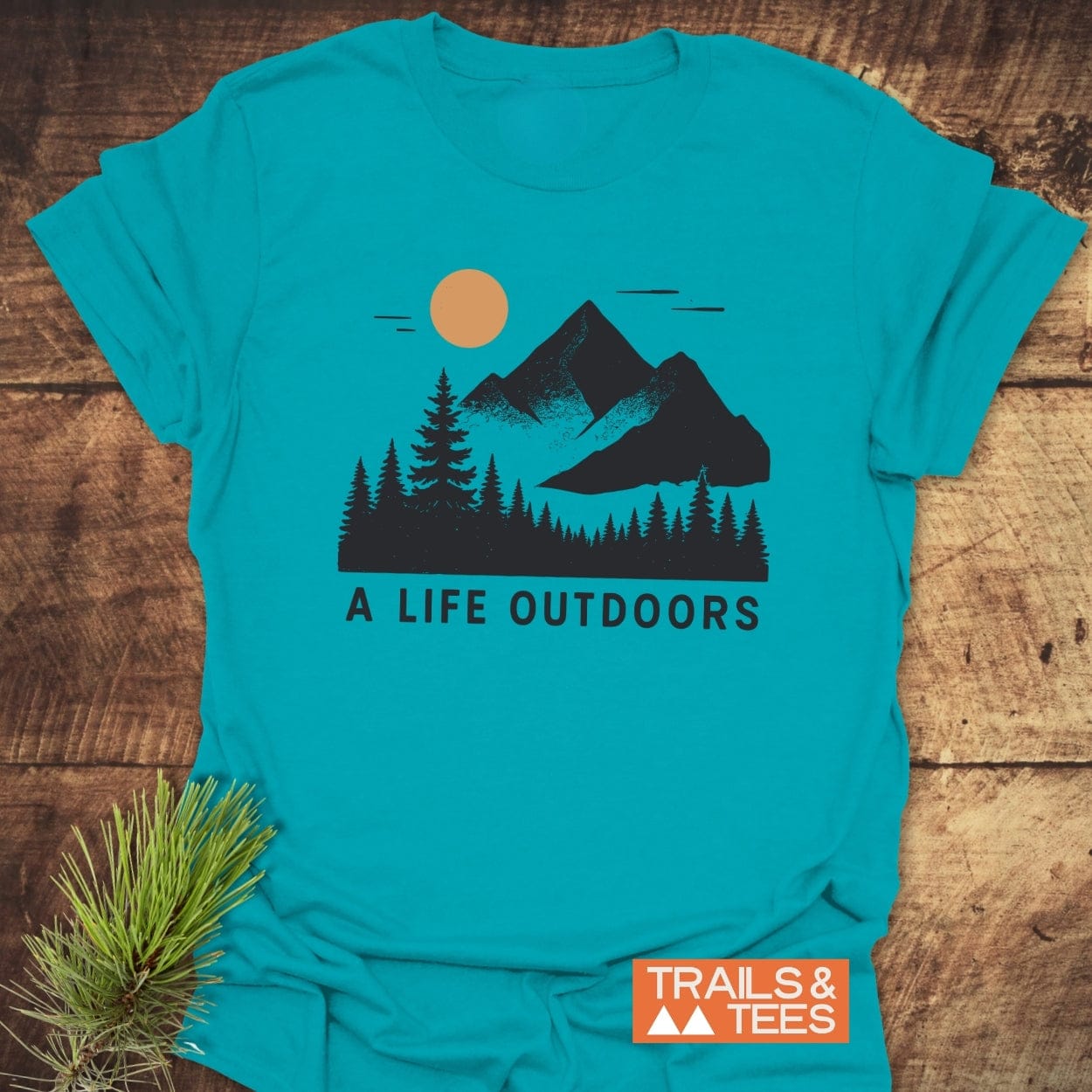 A Life Outdoors T-Shirt with pine trees, mountains, and sun design. Features soft cotton, durable print, and pre-shrunk fit. Perfect for outdoor enthusiasts.
