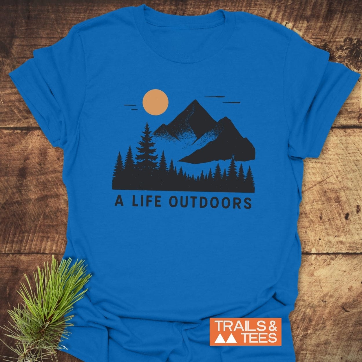A Life Outdoors T-Shirt featuring a stylized outdoor scene with pine trees and mountains, slogan underneath. Made from soft cotton, ideal for everyday adventures.