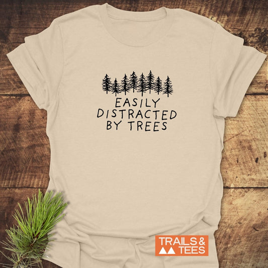 Easily Distracted By Trees T-Shirt with evergreen print, laid on wood. Soft, durable cotton, pre-shrunk, taped shoulders, no side seams. Trails & Tees logo.