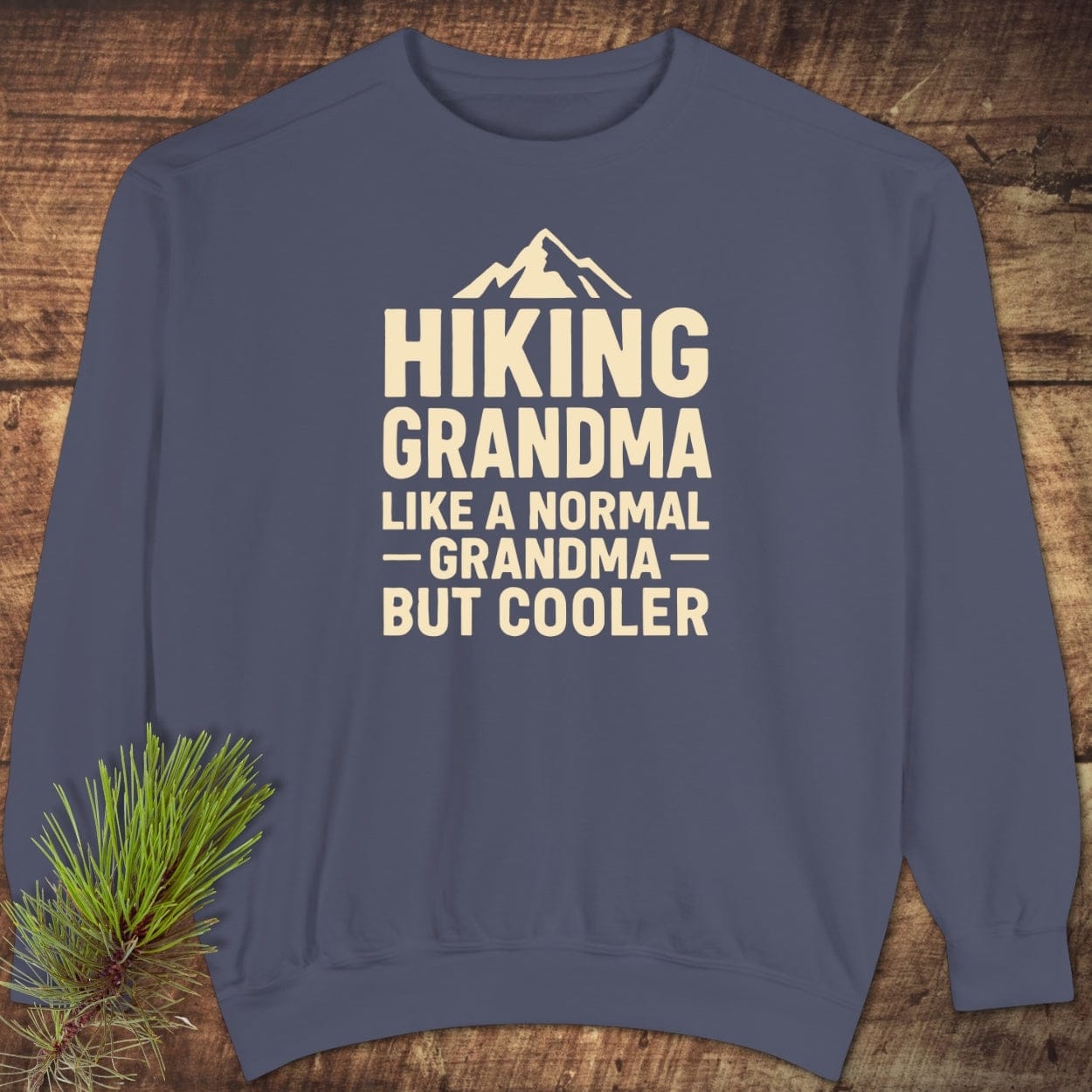 Hiking Grandma Comfort Colors Sweatshirt with mountain graphic; navy-blue, relaxed fit, ring-spun cotton blend, laid out flat on rustic wood background with pine sprig.