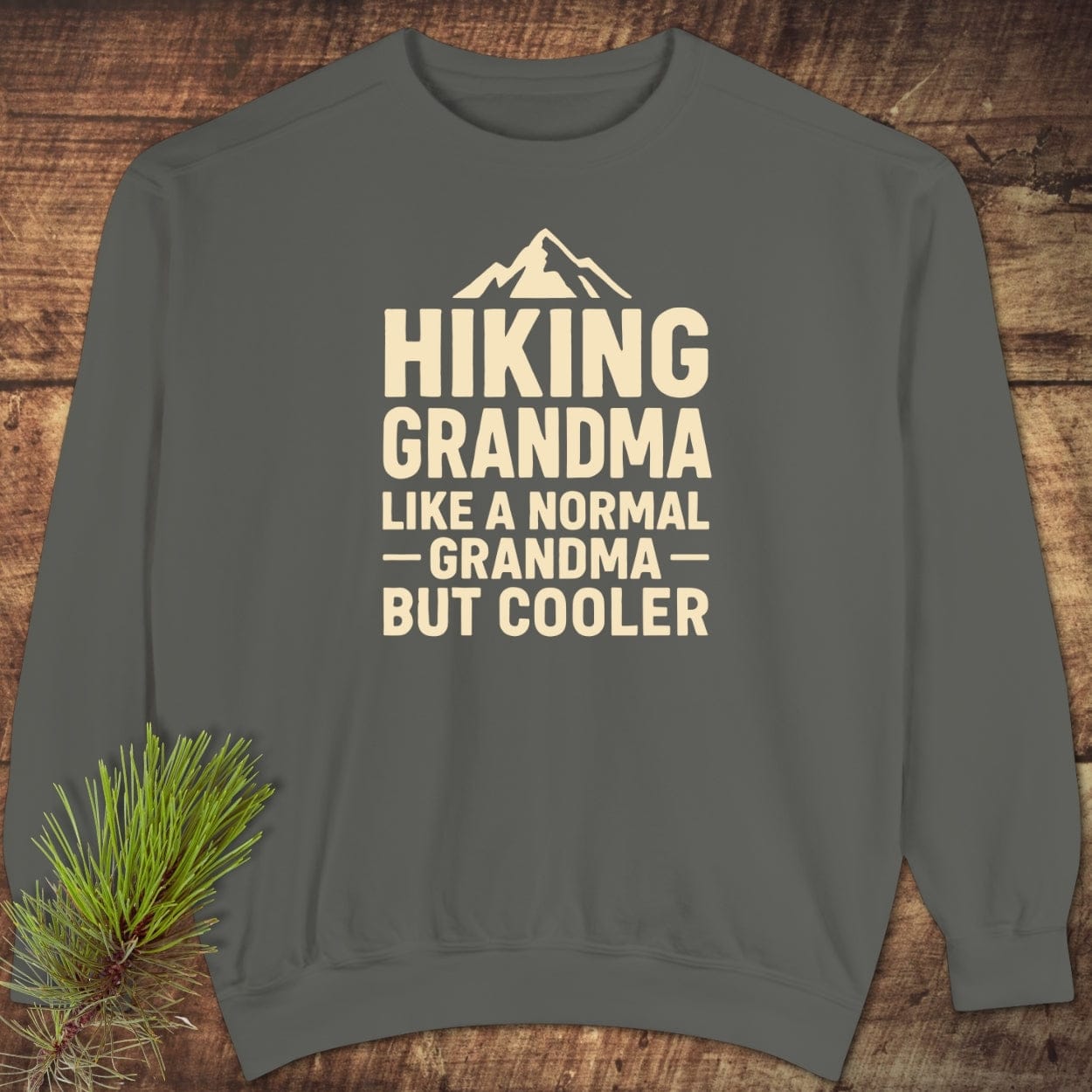 Gray crew-neck sweatshirt with Hiking Grandma Like a Normal Grandma But Cooler text, laid flat on wood, featuring mountain icon. Relaxed fit, unisex, ring-spun cotton blend.