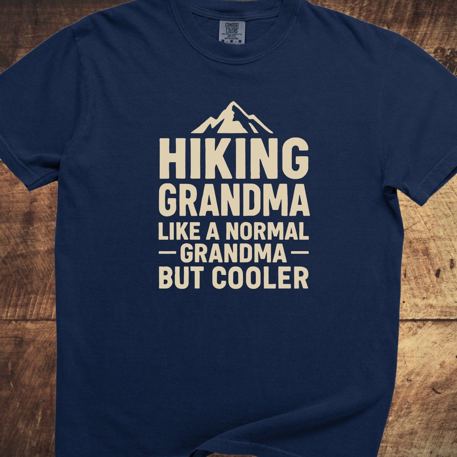Navy-blue Hiking Grandma Comfort Colors 1717 Tee with cream mountain graphic and text, laid flat on wood, showcasing soft-washed, pre-shrunk 100% cotton fabric.