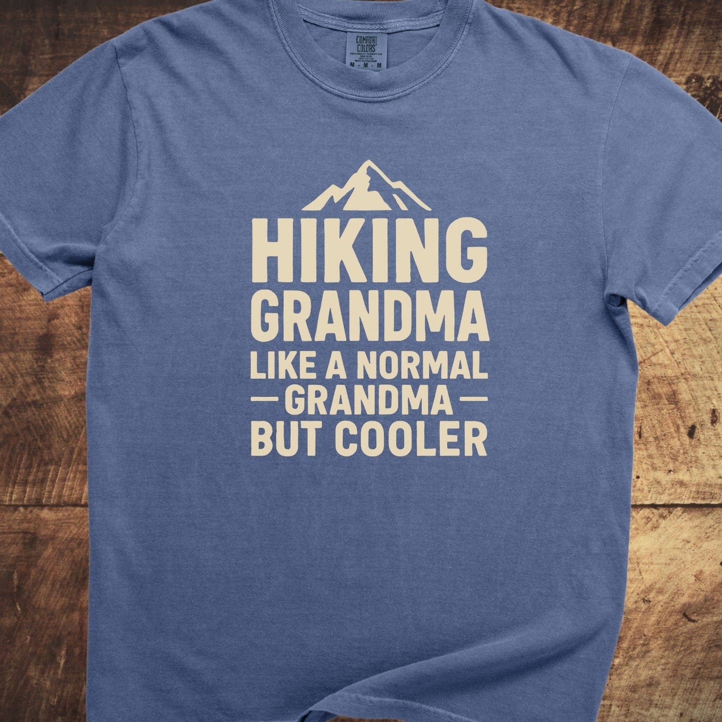Blue Hiking Grandma Comfort Colors 1717 Tee with mountain graphic and playful text, laid flat on wood, showcasing its relaxed fit and durable construction.