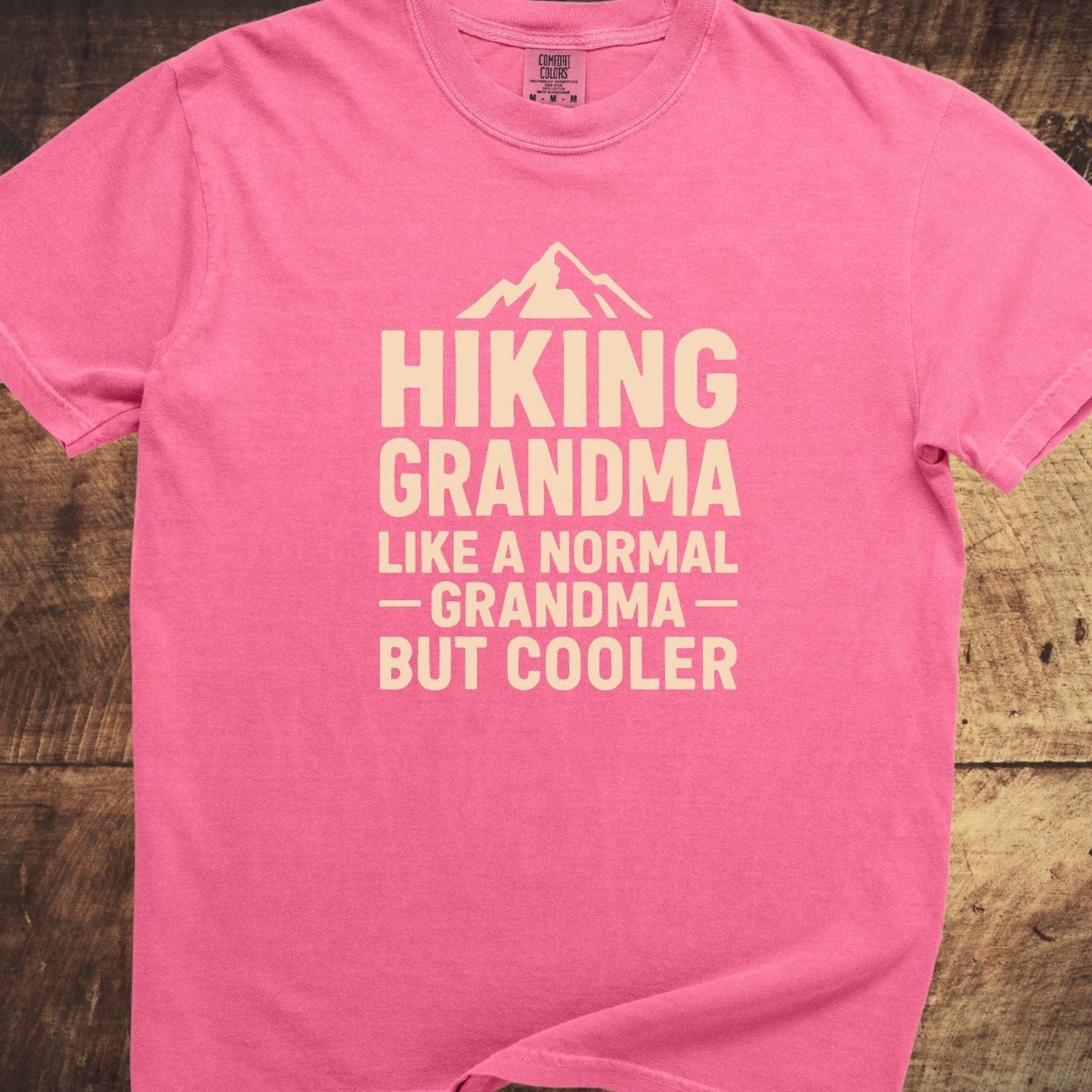 Hiking Grandma Comfort Colors 1717 Tee, featuring a mountain graphic and slogan, laid on wood surface; soft-washed, relaxed fit, durable, pre-shrunk cotton.