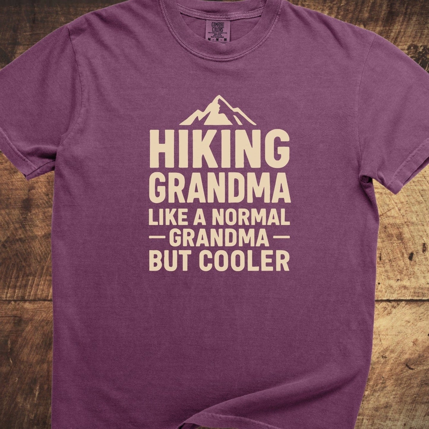 Hiking Grandma Comfort Colors 1717 Tee with mountain graphic and humorous text, laid flat on wood surface, highlighting its relaxed fit and durable design.