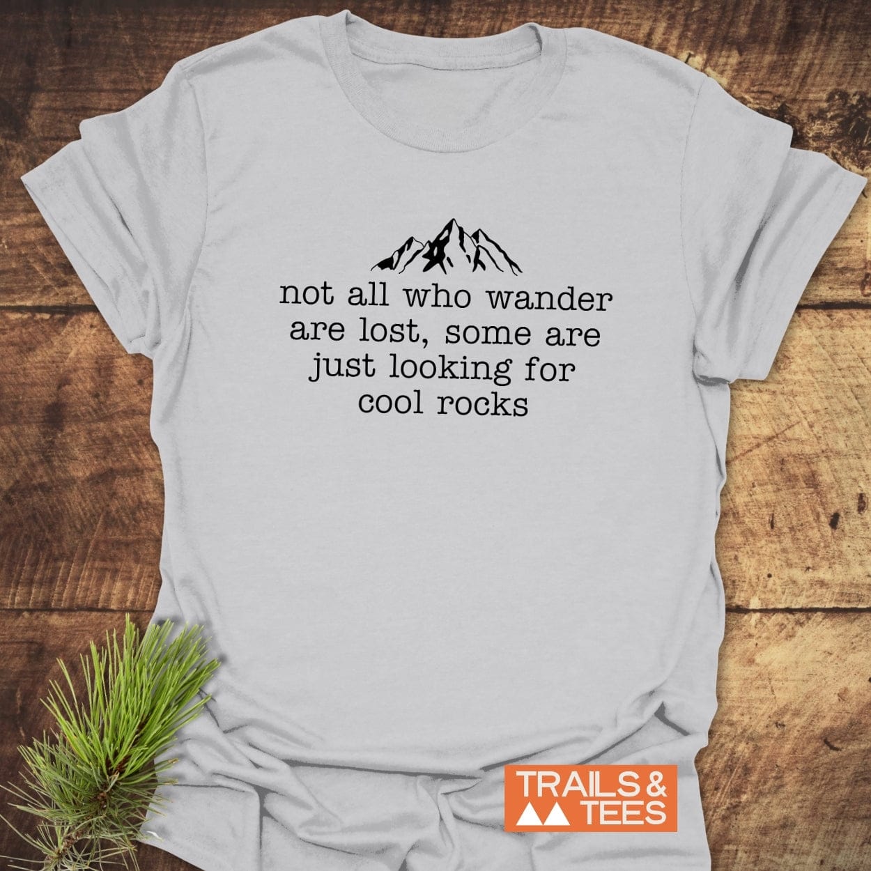 Looking For Cool Rocks T-Shirt featuring a mountain silhouette and quote, laid on wood with pine sprig. TRAILS & TEES. logo in the corner.