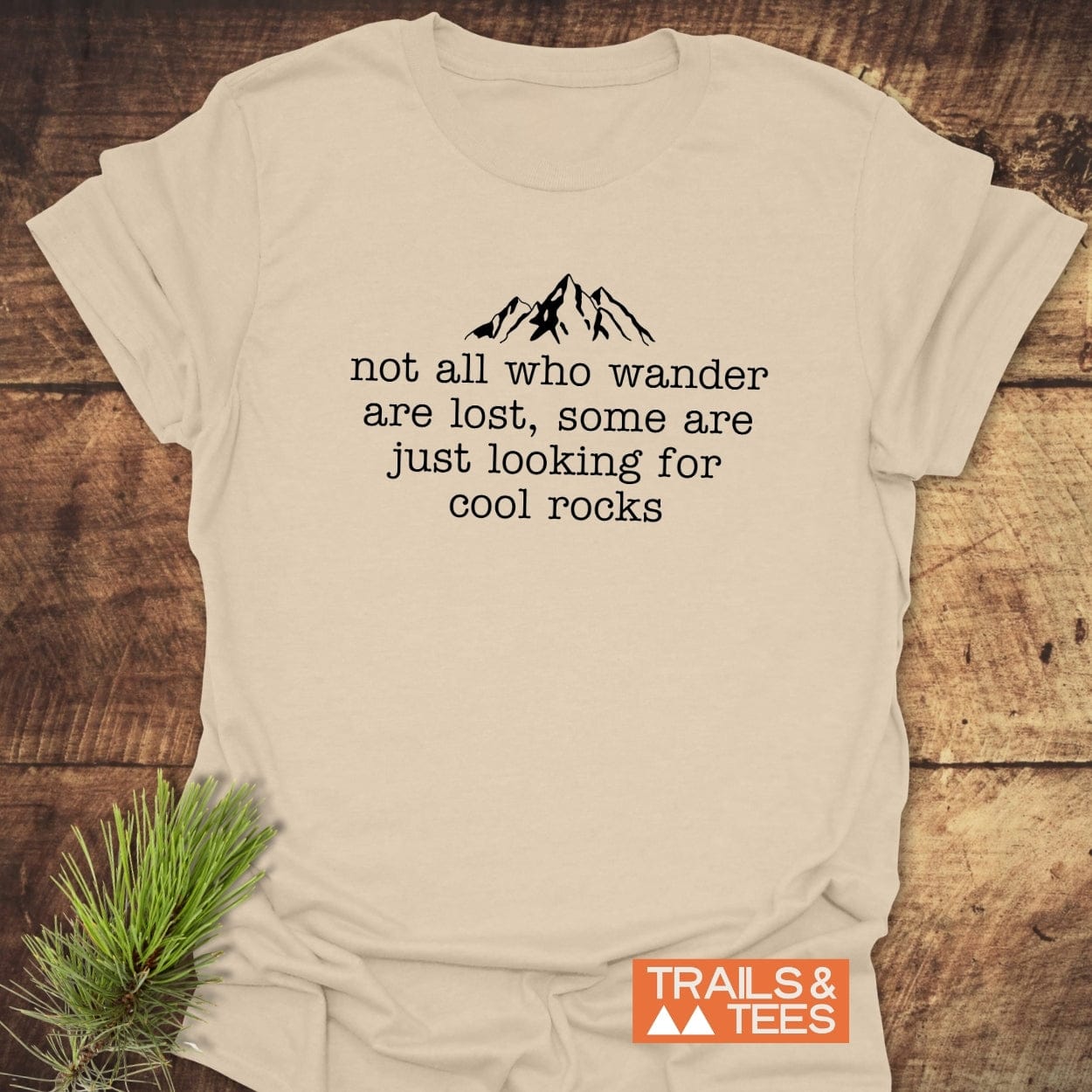 Looking For Cool Rocks T-Shirt on wooden surface, featuring mountain line-drawing and quote. Includes TRAILS & TEES logo, crafted from soft, durable cotton.