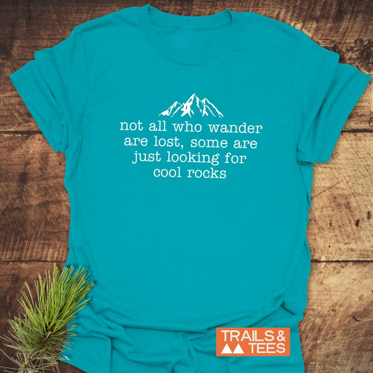 Turquoise-blue T-shirt with mountain silhouette and not all who wander are lost, some are just looking for cool rocks text, by TRAILS & TEES.