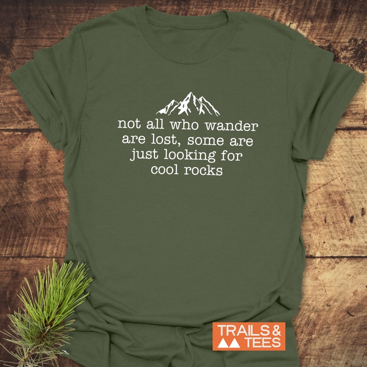 Looking For Cool Rocks T-Shirt, featuring a mountain silhouette and quote, on a wooden surface. Made from soft ring-spun cotton by TRAILS & TEES.