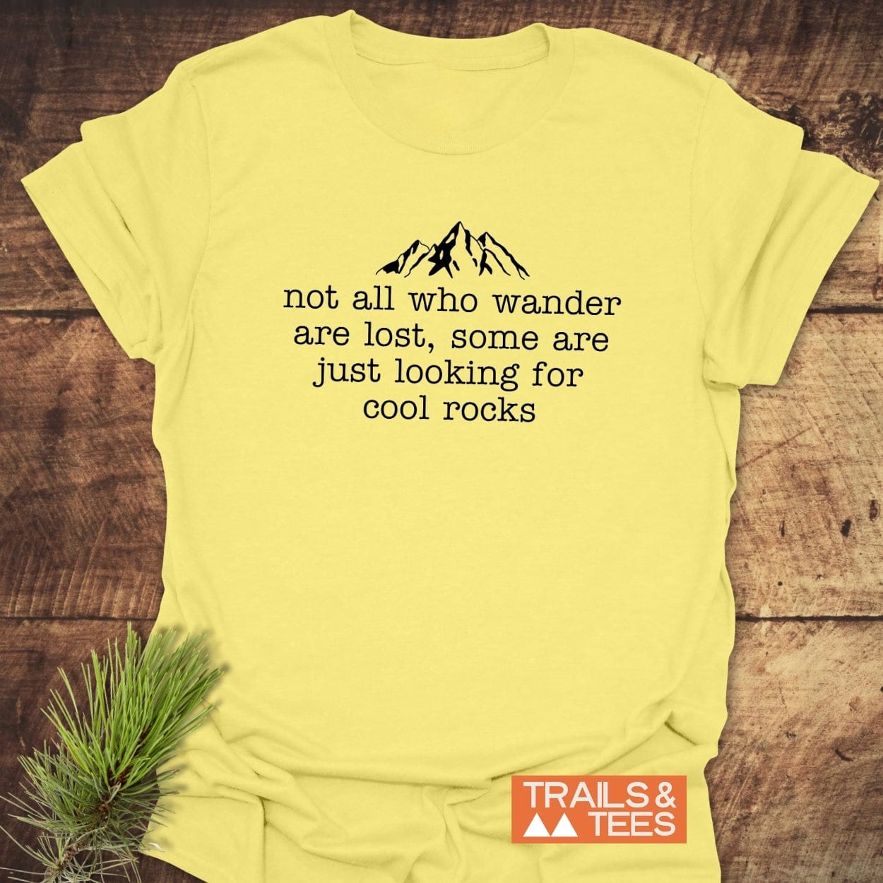 Pale-yellow T-shirt with mountain outline and slogan “not all who wander are lost…” showcasing the Looking For Cool Rocks T-Shirt by TRAILS & TEES.