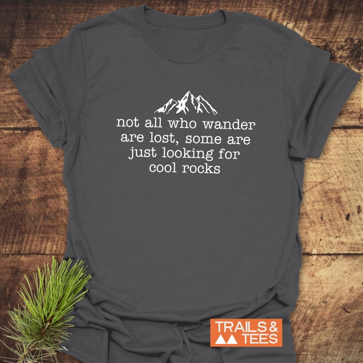 Looking For Cool Rocks T-Shirt with mountain graphic and quote, featuring soft, ring-spun cotton for comfort, laid on a wooden surface with TRAILS & TEES logo.
