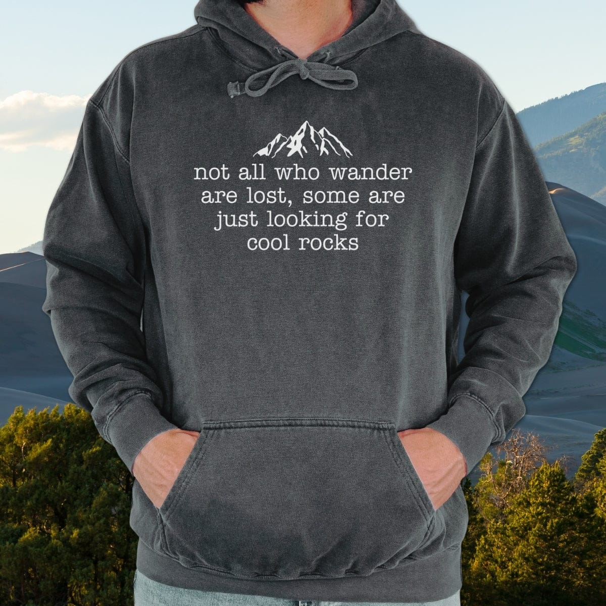 Person wearing the Looking For Cool Rocks Comfort Colors Hoodie with mountain graphic and quote, hands in front pocket, outdoors. Garment-dyed, unisex fit for layering.