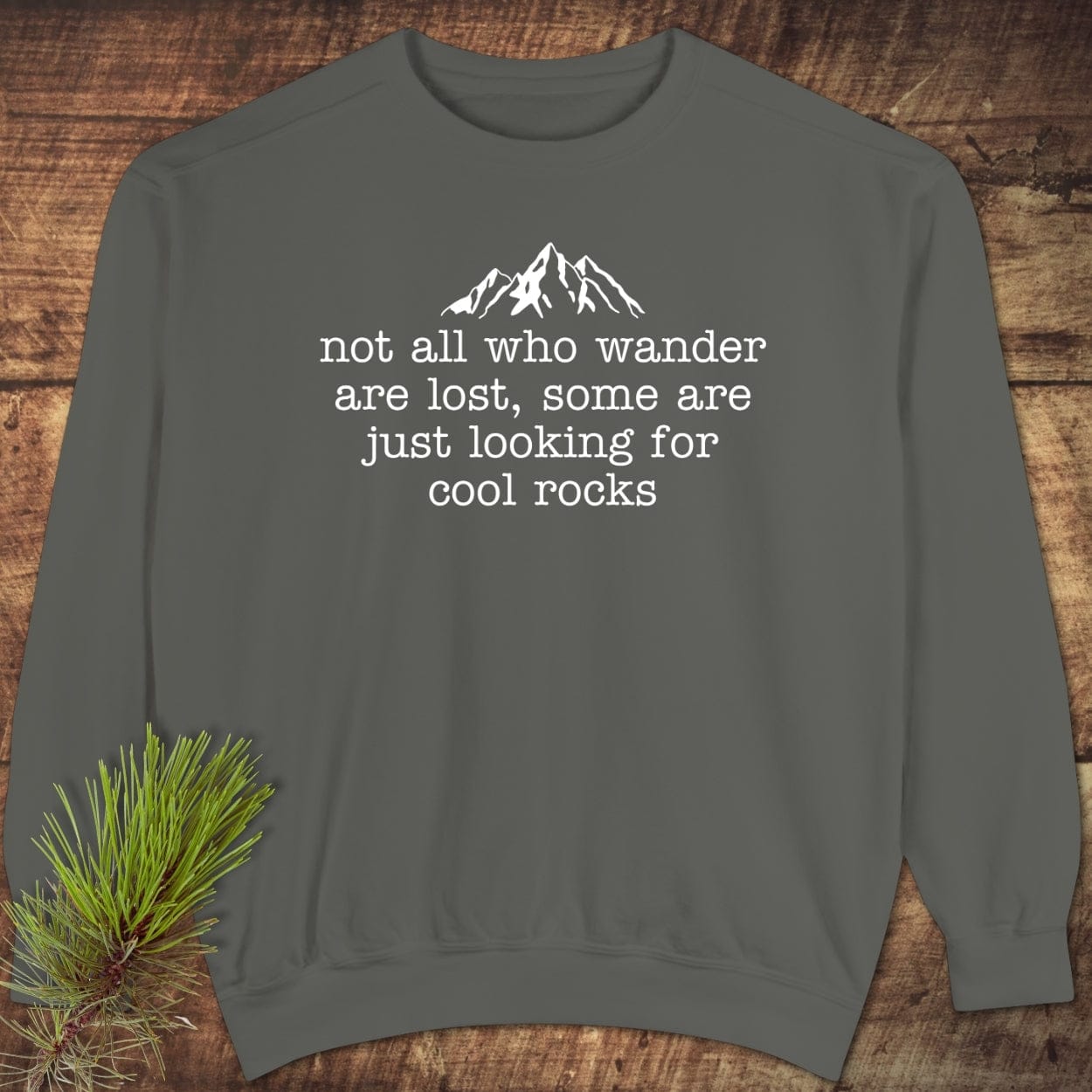 Looking For Cool Rocks Comfort Colors Sweatshirt flat-lay featuring a mountain graphic and quote, styled with a pine sprig on a wooden surface.