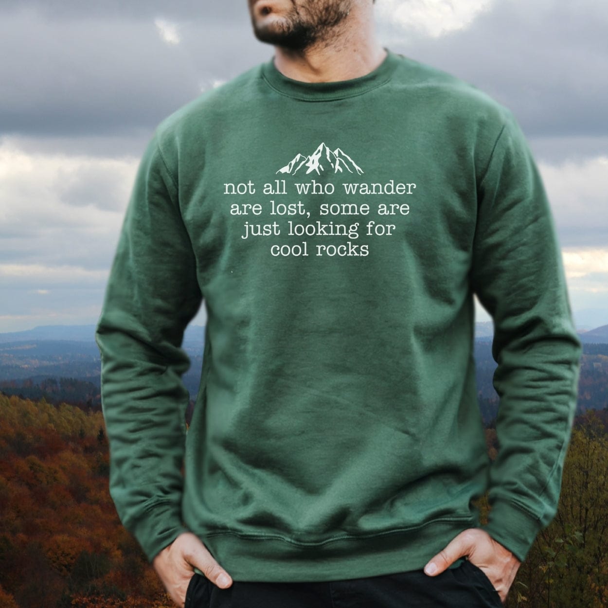 Man wearing Looking For Cool Rocks Comfort Colors Sweatshirt with mountain graphic and quote, showcasing its relaxed fit and luxurious material against an autumn hillside.