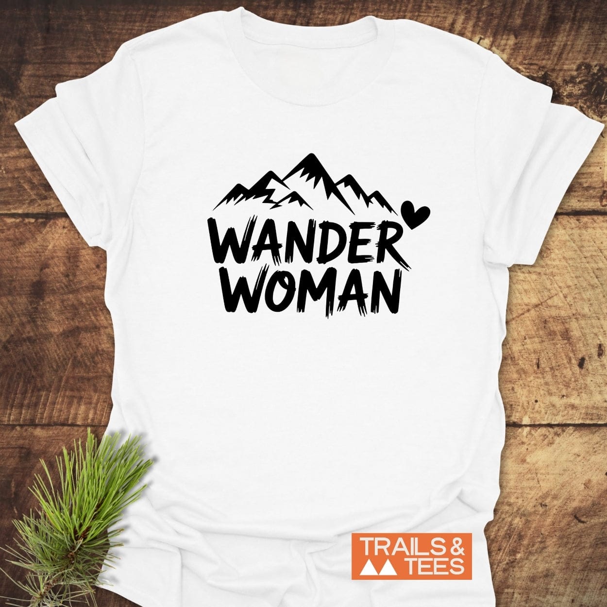 Wander Woman T-Shirt featuring bold black text and mountain silhouette, laid flat with pine needles. Made from soft cotton, durable, pre-shrunk, and adventure-ready.