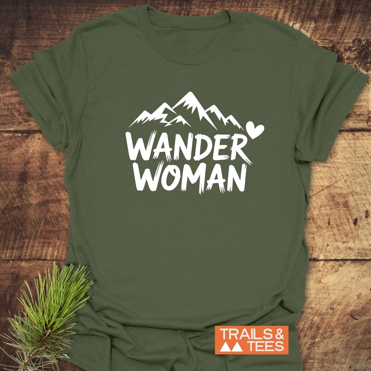 Wander Woman T-Shirt laid on wood with mountain graphic and slogan, featuring soft cotton for comfort, durable print, and nature-themed display with pine sprig.
