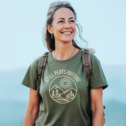 Woman smiling outdoors, wearing Still Plays Outside T-shirt with outdoorsy graphic, showcasing durable, soft cotton ideal for adventure and everyday comfort.