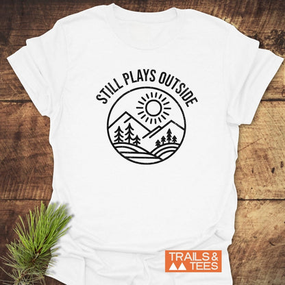 Still Plays Outside T-Shirt featuring a sun, mountains, and trees graphic. Made from soft, lightweight cotton, designed for comfort and durability by Trails & Tees.