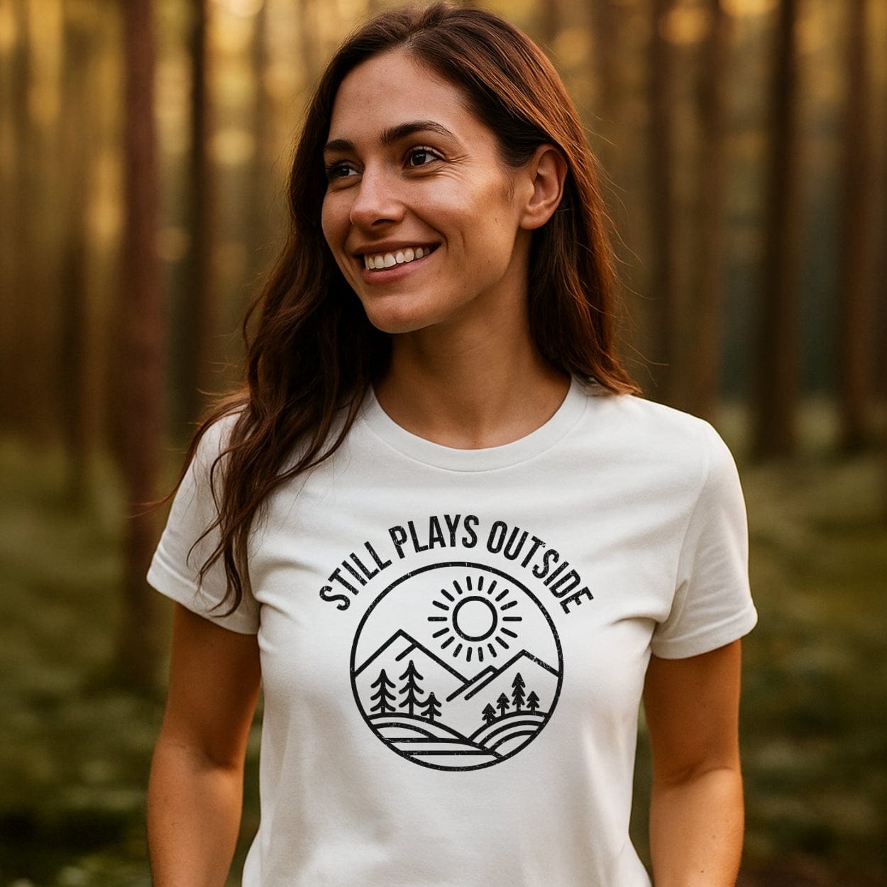Young woman in forest wearing Still Plays Outside T-shirt, featuring mountain and sun graphic, symbolizing trail-tested comfort and outdoor enthusiasm.