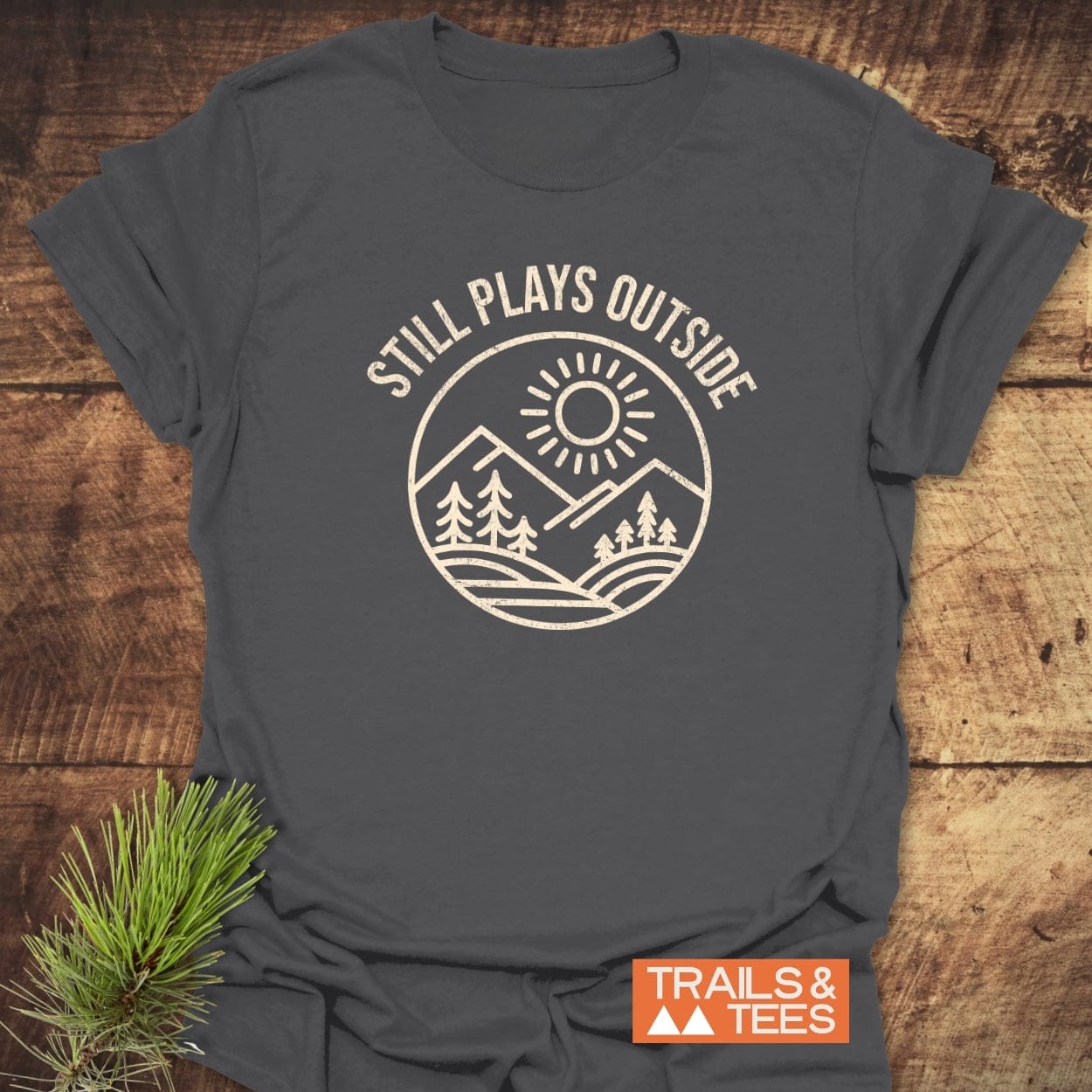 Still Plays Outside T-Shirt: Charcoal-gray tee with outdoor-themed design, ring-spun cotton for comfort, pre-shrunk, durable print, styled with pine needle sprig.