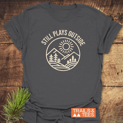 Still Plays Outside T-Shirt: Charcoal-gray tee with outdoor-themed design, ring-spun cotton for comfort, pre-shrunk, durable print, styled with pine needle sprig.