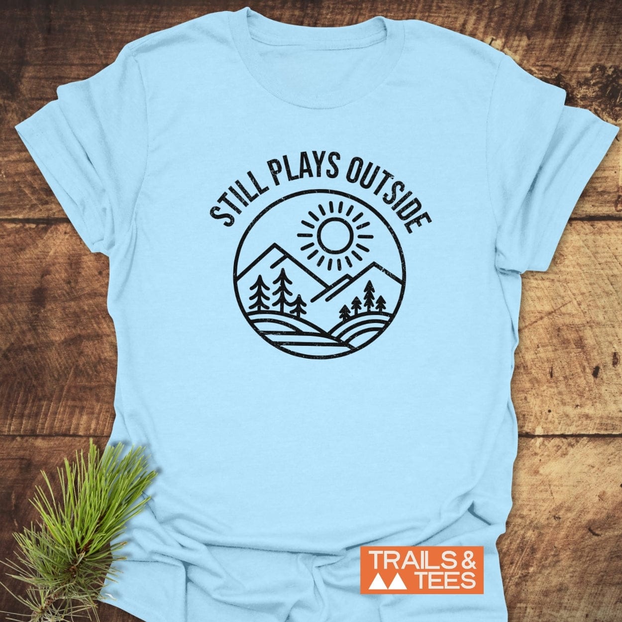 Still Plays Outside T-Shirt featuring a mountain landscape design, crafted from soft ring-spun cotton for comfort and durability, with an orange Trails & Tees label.