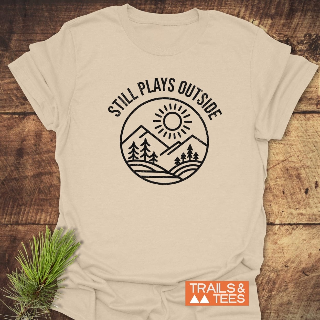 Light-beige Still Plays Outside T-shirt with sun, mountains, and trees design, featuring TRAILS & TEES logo, laid on rustic wood with pine sprig.