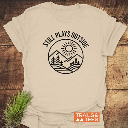 Light-beige Still Plays Outside T-shirt with sun, mountains, and trees design, featuring TRAILS & TEES logo, laid on rustic wood with pine sprig.