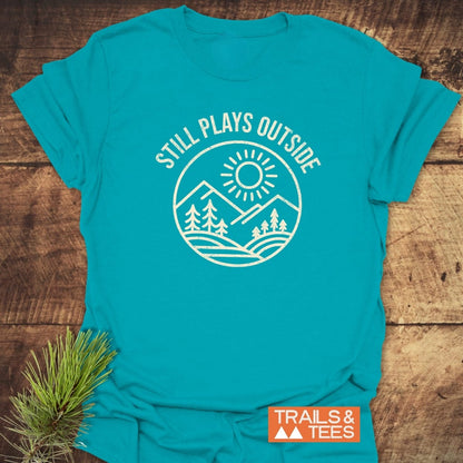 Still Plays Outside T-Shirt on wooden surface, featuring mountain badge and slogan. Includes Trails & Tees logo, pine needles accent, highlighting comfort and adventure-ready design.