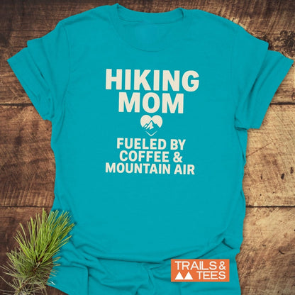 Hiking Mom Mountain Air T-Shirt in teal-blue, features bold white text with a heart-shaped mountain icon, laid on rustic wood with pine sprig styling.