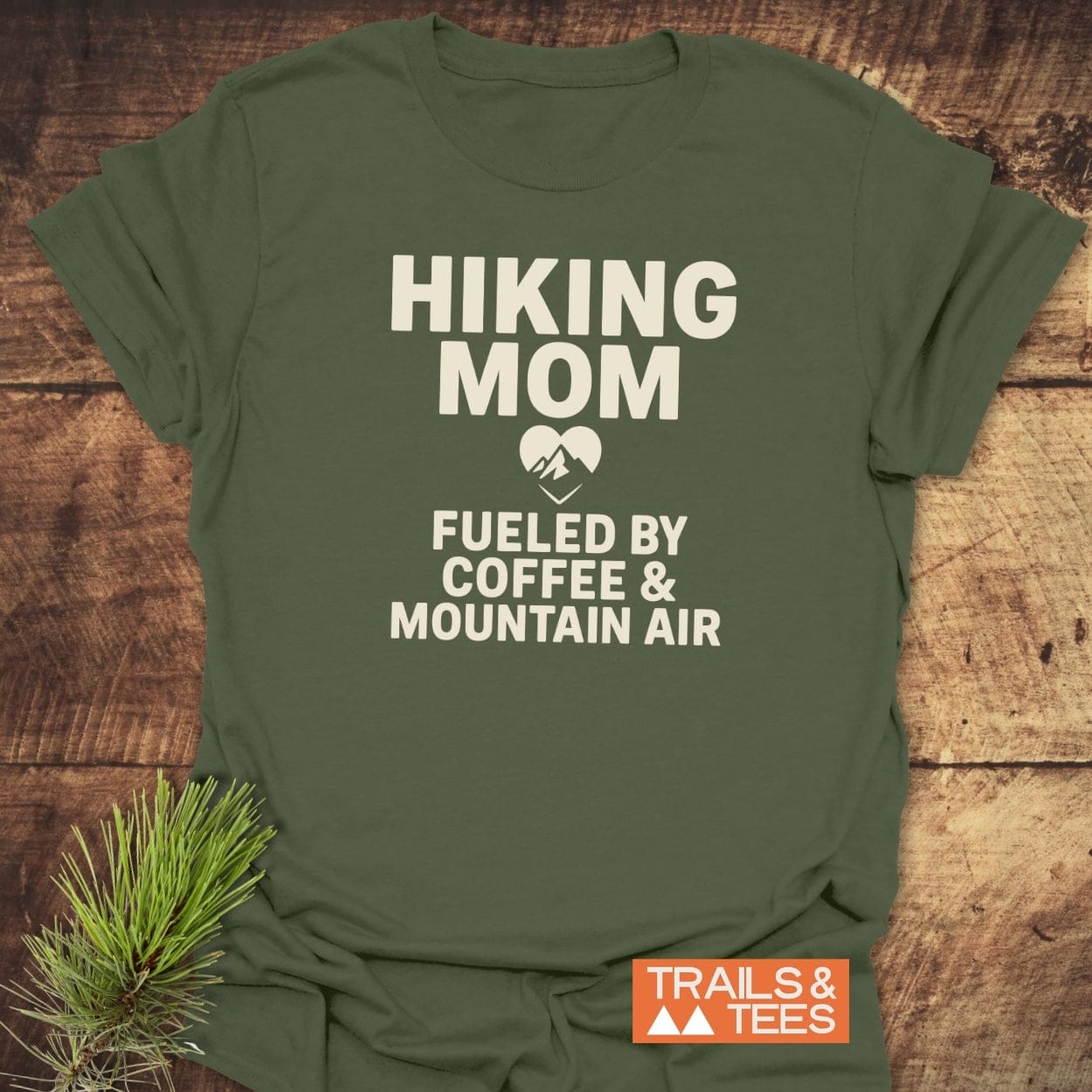 Hiking Mom Mountain Air T-Shirt featuring HIKING MOM and FUELED BY COFFEE & MOUNTAIN AIR prints, styled with a pine sprig on a wooden surface.